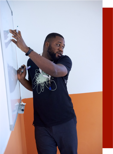 This image shows a man from Ghana standing infront of a whiteboard and explaining what he wrote